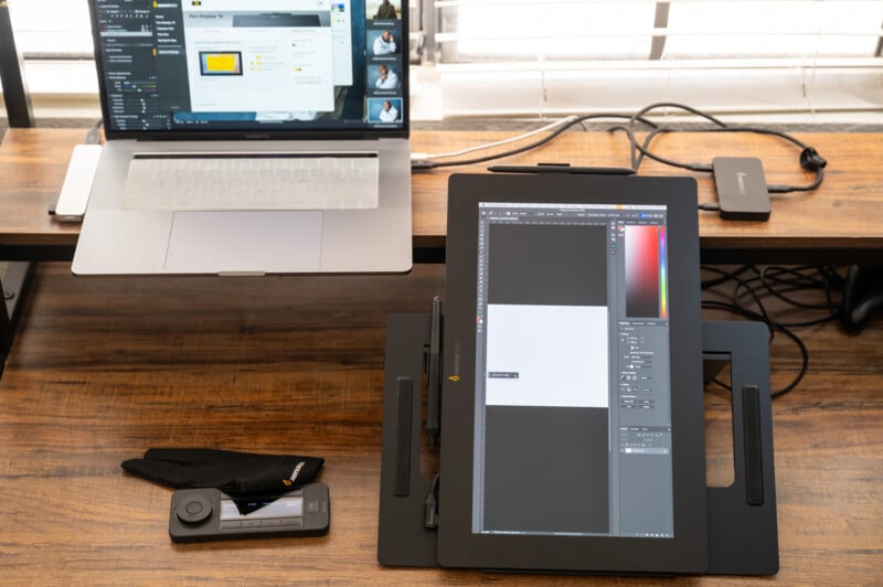 A workspace featuring a laptop and a digital drawing tablet on a wooden desk. The laptop screen shows a design software interface, while the drawing tablet displays an image editing program. Additional accessories include a stylus, a glove, and a color calibration tool.