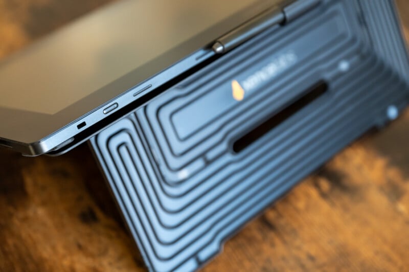 Close-up of a tablet with a durable case, partially propped open on a wooden surface. The tablet's side buttons and part of the screen are visible. The case has a textured, geometric pattern and a logo resembling a flame.