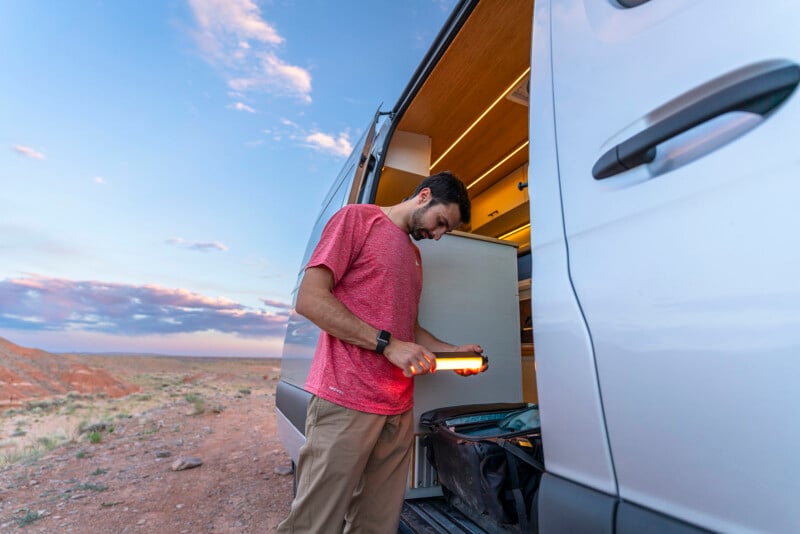 A man wearing a red shirt and khaki pants stands by an open van door. He is holding a light and looking into a bag inside the van. The background features a vast desert landscape and a colorful sky with clouds.