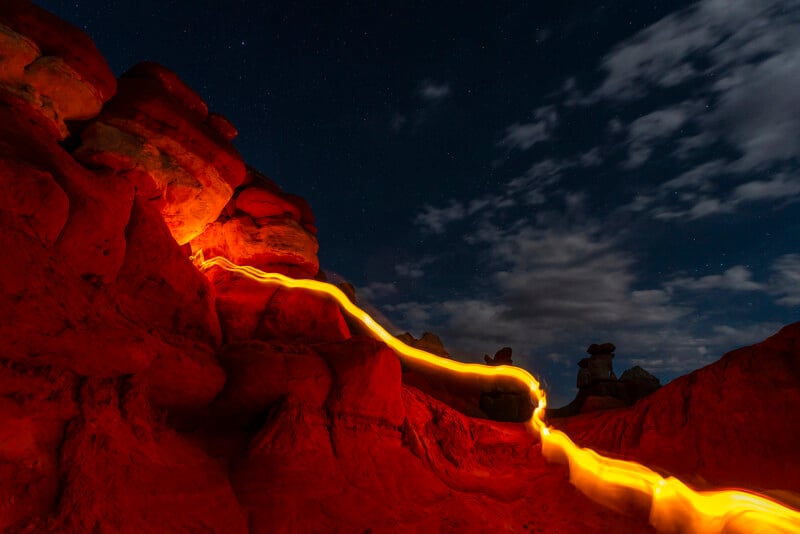 A night sky with stars over a rugged, red rock landscape is illuminated by a wavy trail of yellow light. The formation includes unique rock shapes and shadows, with clouds partially covering the sky.