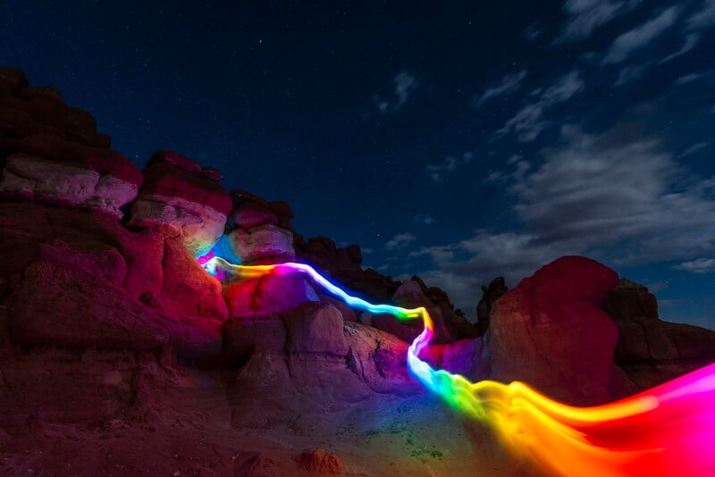 A dramatic night scene in a rocky desert landscape, illuminated by a captivating, winding streak of vibrant light in rainbow colors. The night sky is filled with stars, and some clouds are visible. The rocky formations are partially bathed in colorful lights.