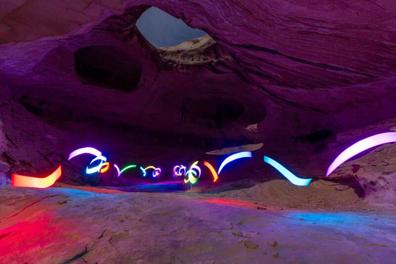A vibrant scene inside a cavern with surreal, colorful light trails weaving throughout. The cave walls shimmer in hues of purple, blue, and red, creating an ethereal atmosphere. The ceiling features a large, natural hole, allowing a hint of the night sky to peek through.