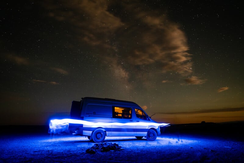 A van parked in a desert under a clear starry night sky. The van is partially illuminated with blue light trails, creating a swirling effect around it. A campfire ring made of rocks is in the foreground, and wispy clouds are visible against the starry backdrop.