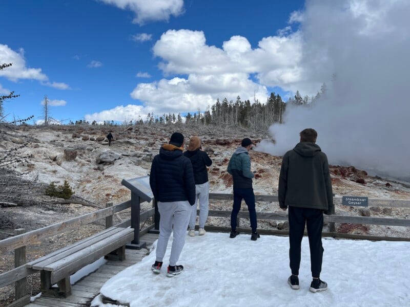 Four people in winter clothing are facing away from the camera, observing the Steamboat Geyser at Yellowstone National Park. Steam is rising from the geyser, and the ground is covered in snow. The sky is partly cloudy, and trees line the background.