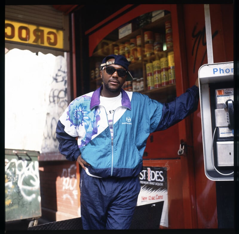 A person wearing a blue and white tracksuit, black sunglasses, and a headband stands next to a payphone. They are posed casually with one hand on their hip in front of a storefront with visible graffiti and goods displayed in the window.