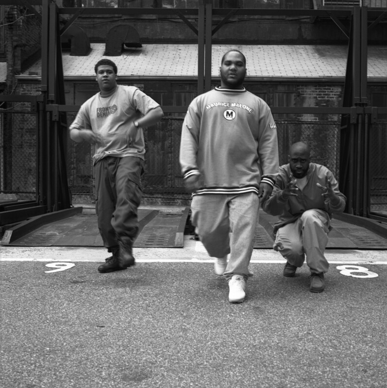 Three men are walking and posing on the street in front of a multi-level car parking structure. The man on the left is wearing a sweatshirt, the man in the middle has a large jacket, and the man on the right is crouching and wearing a jacket and pants. The photo is black and white.