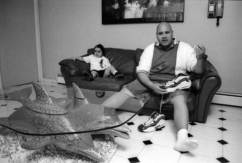 A man sits on a couch holding a sneaker with another on his foot. He's in a casual outfit and appears to be talking. A child is seated on another couch. The room has a unique dolphin-shaped glass table and a painting on the wall in the background.