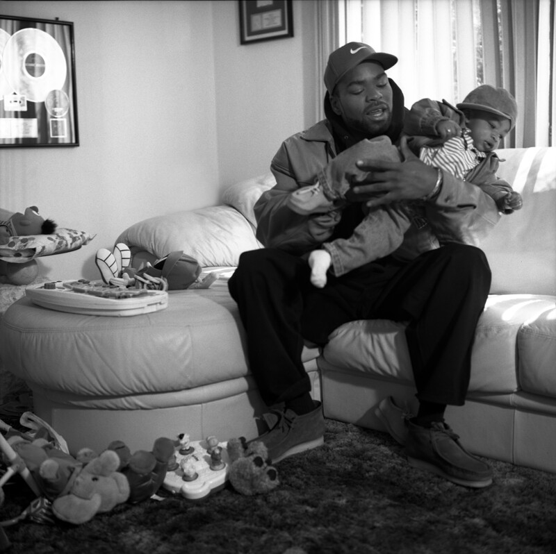 A man wearing a cap sits on a couch holding a baby dressed in a hat and striped outfit. The living room has toys scattered on the floor and couches, including stuffed animals and a playset. Sunlight streams in through a window with curtains in the background.