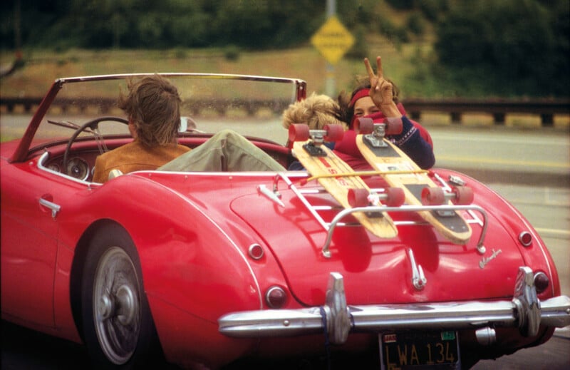 A bright red convertible with two longboards strapped to the luggage rack speeds along a highway. Two passengers, one showing a peace sign, smile and enjoy the ride with the wind in their hair. The car's top is down offering a clear view of the scenic surroundings.