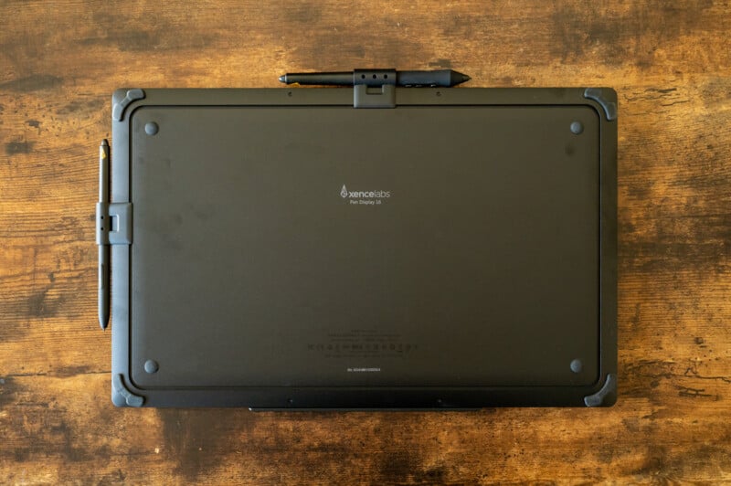 A top-down view of a black rugged tablet with a stylus attached to the top. The tablet rests on a wooden surface, and the brand name "Xplore" and model info are visible on the back. Corners are reinforced with protective bumpers.