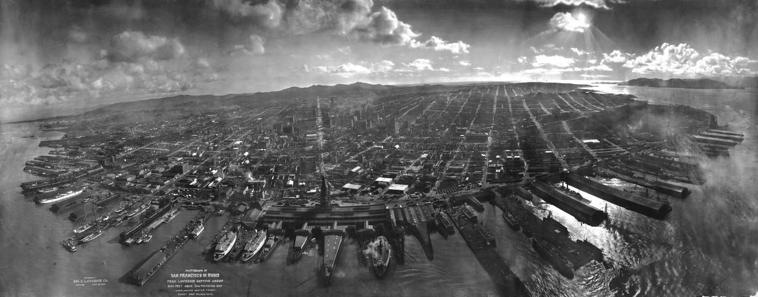 A grayscale aerial panorama of San Francisco, captured in 1920. The image shows the city's extensive waterfront, numerous docks, and ships along the bay. The urban landscape stretches back towards rolling hills under a partly cloudy sky.