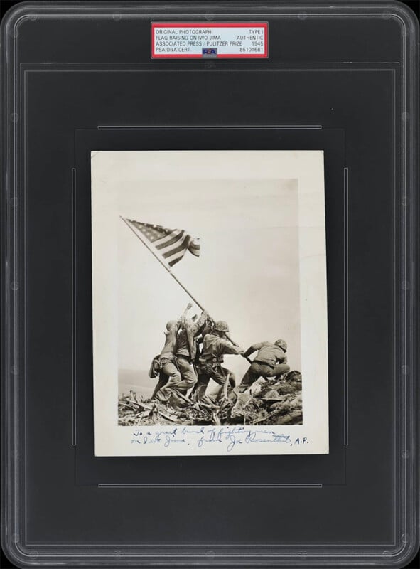 A framed and certified photograph of U.S. soldiers raising the American flag on Iwo Jima during World War II. The black-and-white image shows six soldiers working together to plant the flag on a rocky terrain. There is handwritten text at the bottom of the photo.