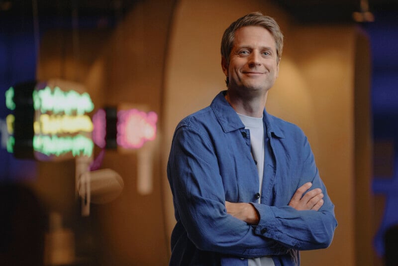 A man with short blonde hair and a light beard stands confidently with arms crossed. He is wearing a blue button-up shirt over a white T-shirt, smiling softly, with a blurred, warmly lit background featuring colorful, neon-like light fixtures.