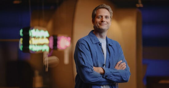 A person with short hair stands confidently with arms crossed. They are wearing a blue button-up shirt and smiling slightly. The background features neon lights with various colors, creating a modern and artistic ambiance.