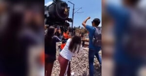 A group of people stands on a gravel surface taking photos and enthusiastically cheering as a vintage steam locomotive passes by. One person is raising their fist in excitement, while others use their phones to capture the moment. The sky is clear and blue.