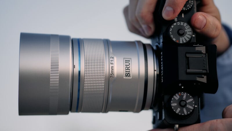 Close-up of hands holding a camera with a silver lens. The lens has "Sirui" and "75mm F1.2" written on it. The camera's dials and buttons are visible, and the background is out of focus.