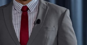 A person in a grey suit jacket, a white shirt with thin black stripes, and a red tie stands in front of a blue curtain. A small black lavalier microphone is clipped to the jacket's lapel. Only the torso and part of the face are visible.