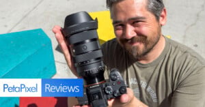 A person with a beard and gray hair smiles while holding a Sony camera with a large lens. The background features colorful geometric shapes, and there is a "PetaPixel Reviews" label in the lower left corner of the image.