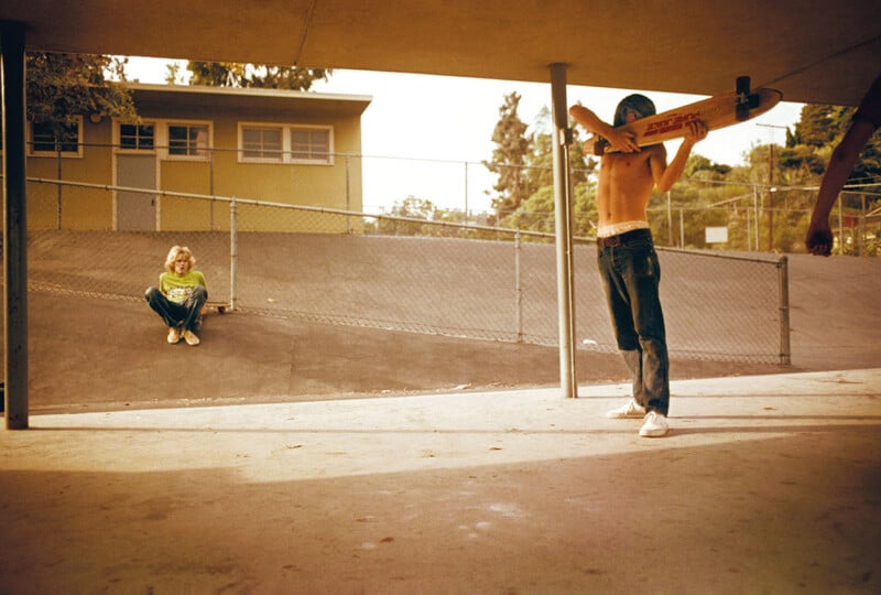 A shirtless person with a skateboard over their shoulder stands under a shaded area, facing away. In the background, a person in a green shirt sits against a fence beside a ramp. The scene is bathed in warm light, suggesting late afternoon.