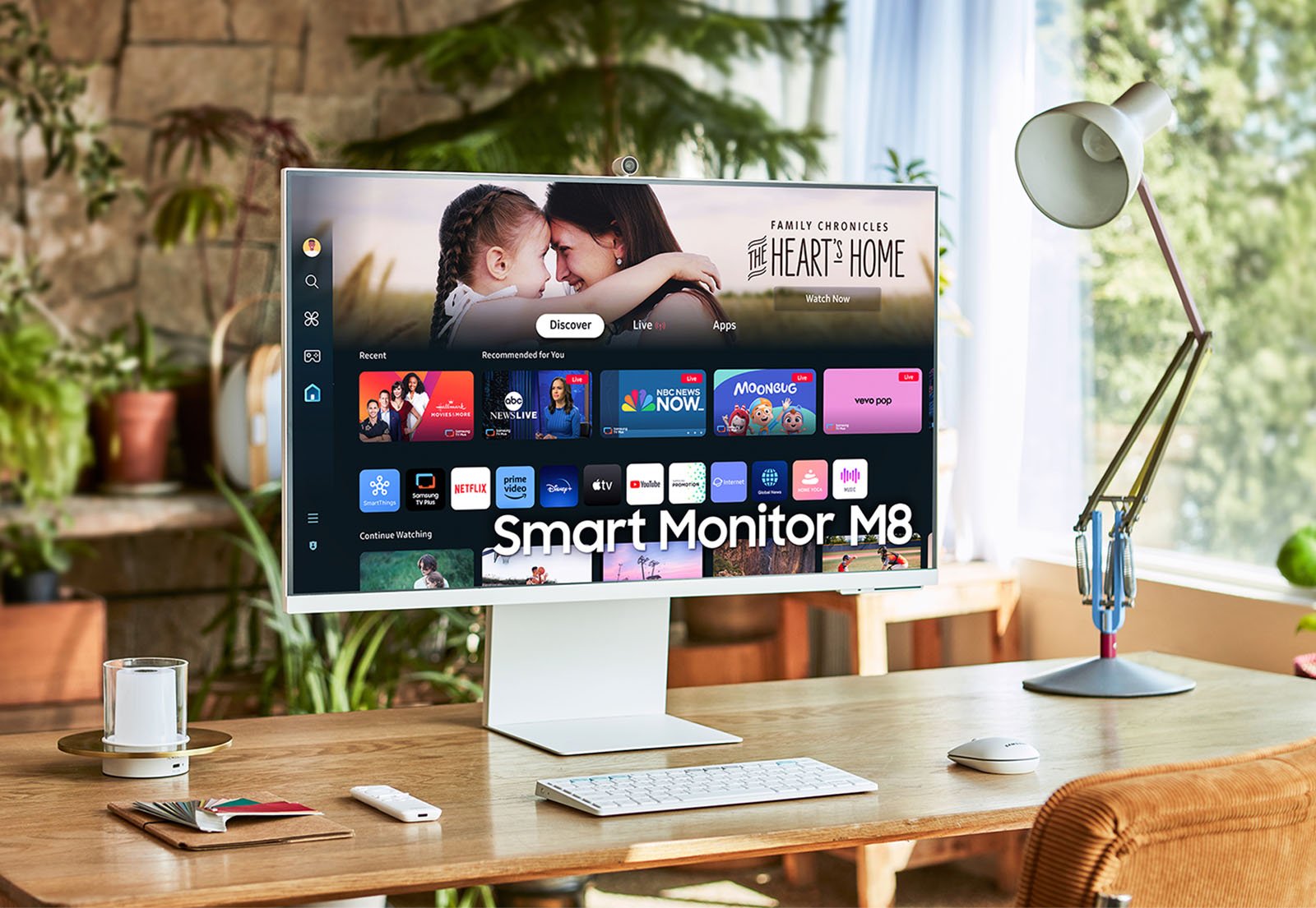 A sleek, modern desk setup featuring the Samsung Smart Monitor M8. The monitor displays a streaming service interface with various entertainment app icons. The desk includes a lamp, cup of coffee, pens, and an open book. The background showcases a cozy, well-lit room.