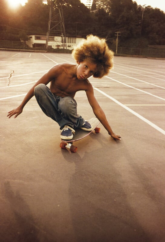 A young person with curly hair skateboards shirtless on an empty asphalt lot. They are balancing on one hand while crouching low on the board, wearing jeans and blue sneakers. Trees and structures are visible in the background under a warm, golden light.
