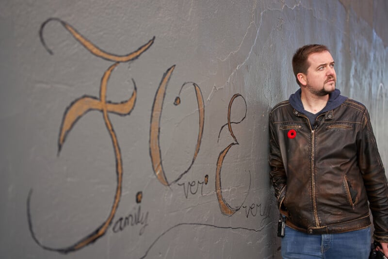 A man in a leather jacket with a red flower pin stands against a graffiti-covered wall. The graffiti includes the word "FOE" in large, stylized letters, with smaller text "family" and "over" visible below it. The man gazes off to the right, hands in his pockets.