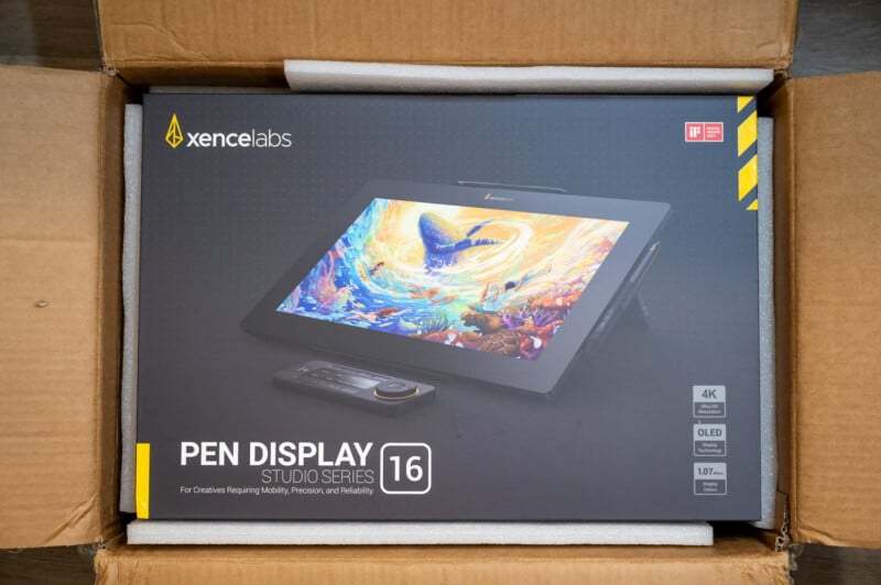 The image shows the packaging of a Xencelabs Pen Display Studio Series 16. The box features a vibrant illustration on the tablet screen, with text highlighting its 4K resolution, OLED display, and various features for creative professionals. The box is inside a cardboard shipping container.