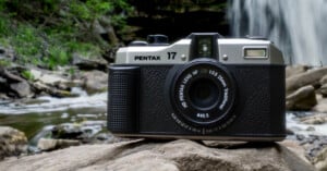 A vintage Pentax 17 camera sits on a rock with a waterfall and lush greenery in the background. The camera is positioned slightly to the right, highlighting its lens and intricate design amidst the natural setting.