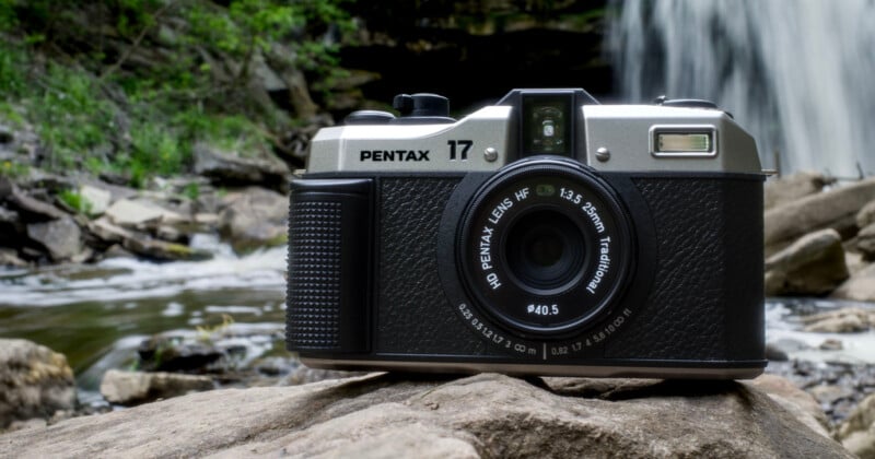 A vintage Pentax 17 camera sits on a rock with a waterfall and lush greenery in the background. The camera is positioned slightly to the right, highlighting its lens and intricate design amidst the natural setting.