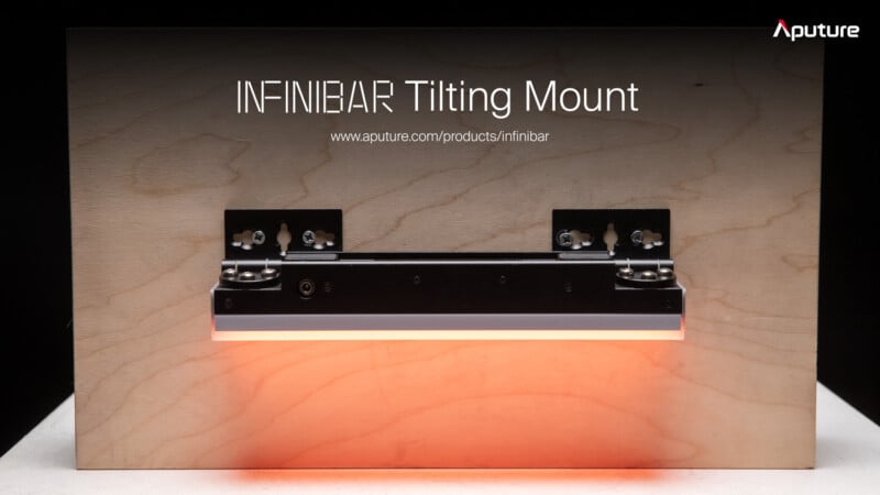 A tilting mount for a light bar is attached to a wooden panel. The text "INFINIBAR Tilting Mount" and a website link "www.aputure.com/products/infinibar" are displayed above the mount. The light bar emits a soft orange glow. The Aputure logo is in the upper-right corner.