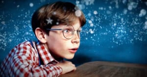 A young boy with glasses and freckles stares in awe at a magical, glittering scene in front of him. He is wearing a red checkered shirt and has his hands resting on a wooden surface. The background is filled with sparkling lights and a blue, starry atmosphere.