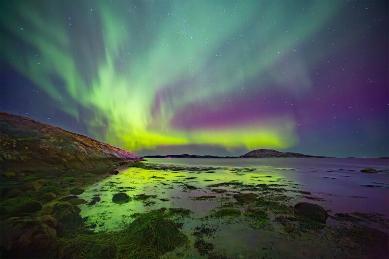 A tranquil shoreline under a starry sky is illuminated by vibrant green and purple hues of the Northern Lights. The vivid aurora reflects off the water and wet stones, creating a mesmerizing and colorful display in the serene natural landscape.