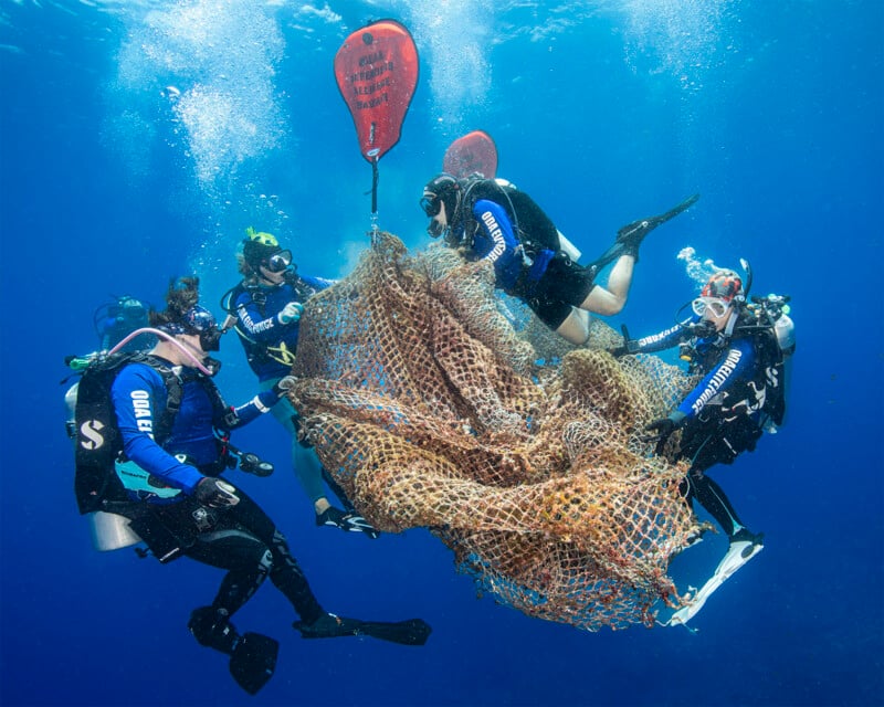 Five scuba divers work together underwater to remove a large fishing net from the ocean. They are wearing full diving gear, including wetsuits and tanks, and are pulling the net toward a red buoyancy bag to bring it to the surface. The water around them is clear and blue.