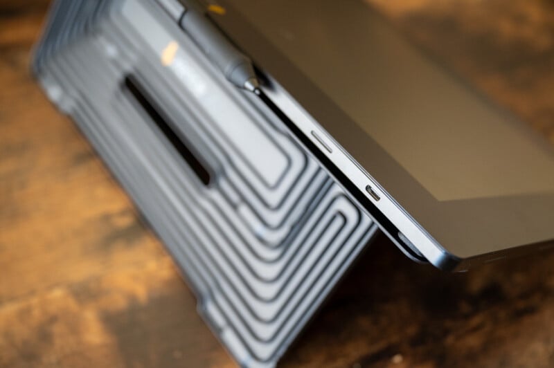 A close-up of a tablet placed on a textured, geometric-patterned stand. The tablet screen is partially visible, with edges reflecting light. A stylus is attached to the stand. The background is a wooden surface that appears slightly blurred.