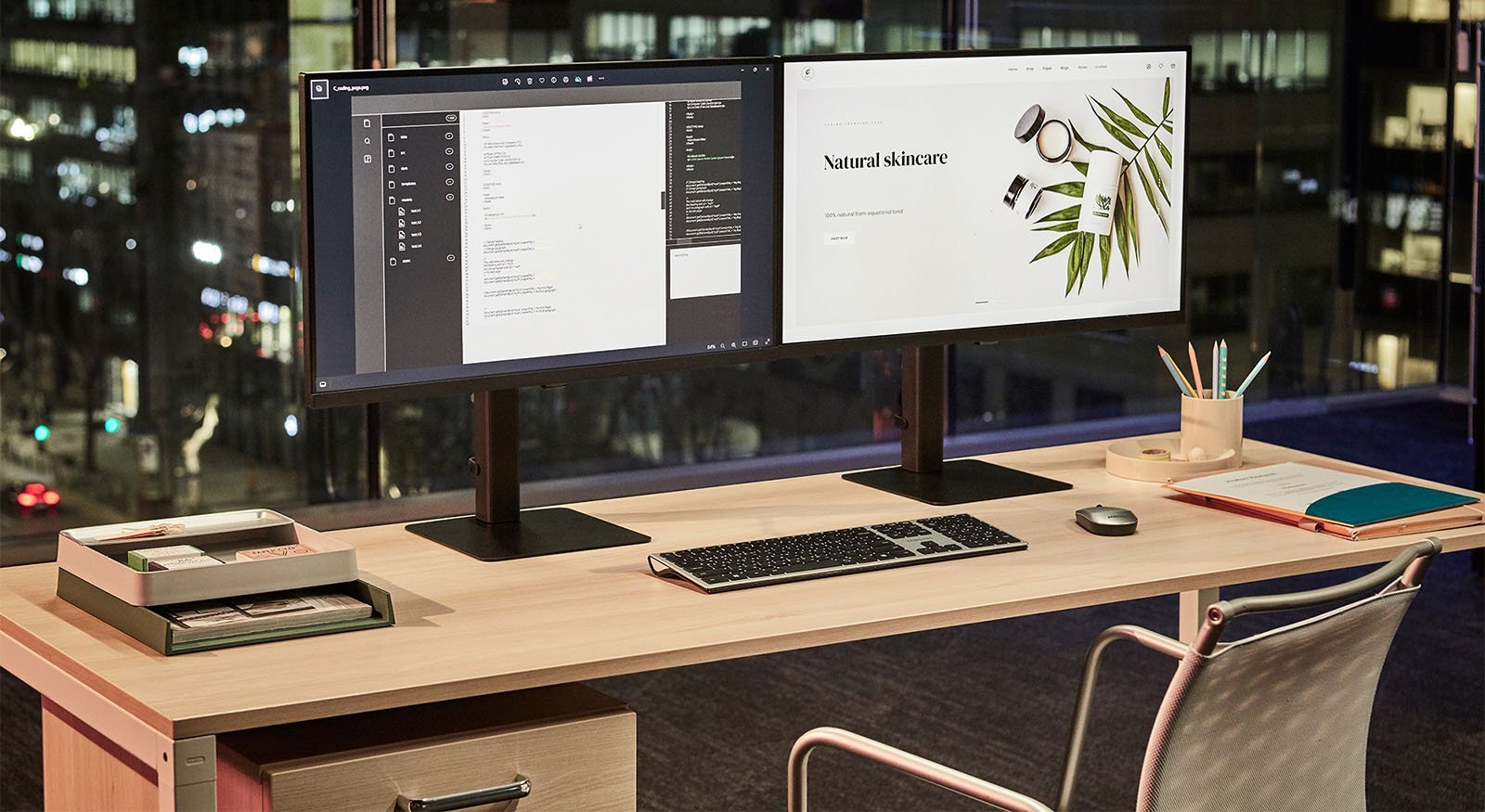 A modern office setup with dual monitors on a desk. One screen displays code, the other shows a natural skincare website. The desk has a keyboard, mouse, notepads, pen holder, and office supplies against a backdrop of city lights through a window.