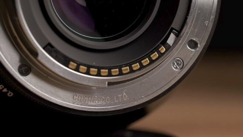 Close-up image of the mount of a camera lens, displaying the electronic contacts and the inscription "COSINA CO., LTD." on the metal surface. The background is blurred, drawing focus to the detailed elements of the lens mount.