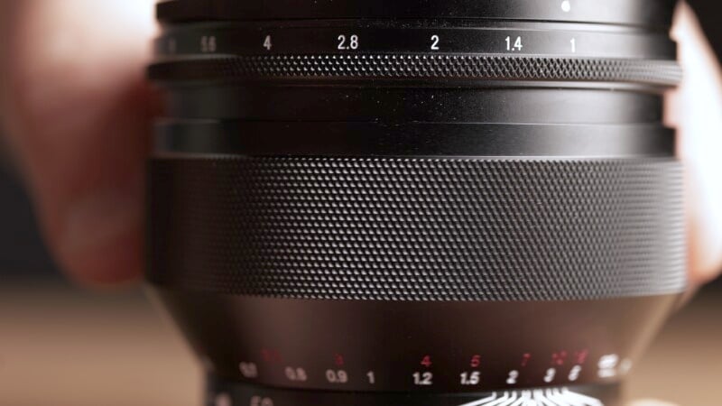 Close-up view of a black camera lens with a focus ring marked with numbers from 1.4 to 2.8 and a textured grip. The aperture ring below it features red and white numeric markings indicating different f-stop values. A hand partially holds the lens.