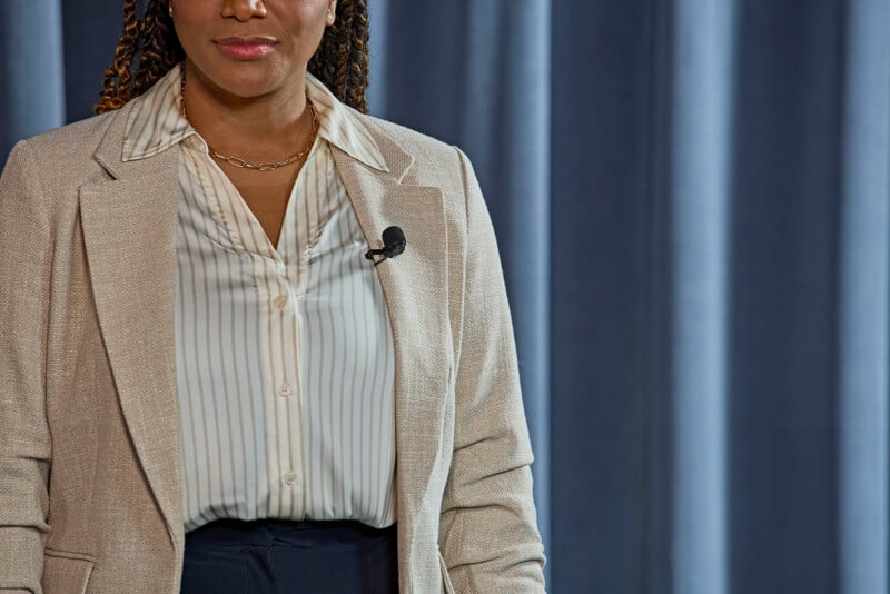 A person wearing a beige blazer over a white striped shirt and a chain necklace is standing in front of a blue curtain. A microphone is clipped to the lapel of their blazer. The image focuses on the person's upper body and does not include the person's full face.