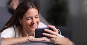 A woman with long brown hair smiles while looking at her smartphone. She is wearing a white shirt and has white earphones in her ears. The background is blurred, focusing attention on her joyful expression.