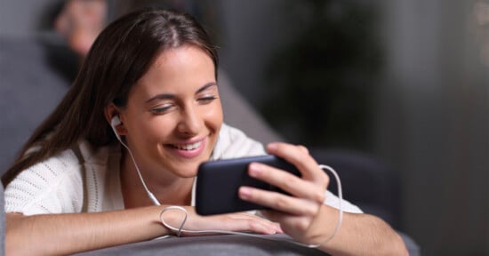 A woman with long brown hair smiles while looking at her smartphone. She is wearing a white shirt and has white earphones in her ears. The background is blurred, focusing attention on her joyful expression.