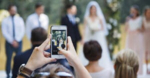 A person holds up a smartphone to take a photo of a bride and groom standing together at an outdoor wedding ceremony. The bride is in a white dress and veil, the groom in a suit. Blurred guests and greenery are visible in the background.