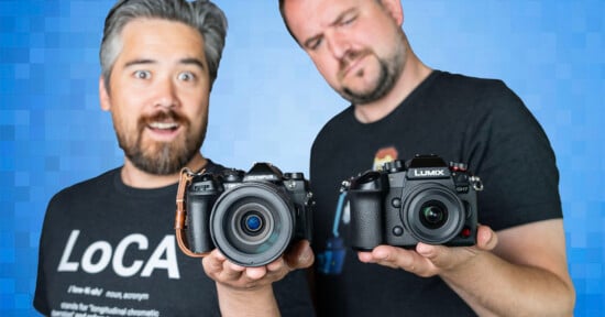 Two men are holding cameras towards the viewer, smiling and standing against a blue pixelated background. The man on the left holds an Olympus camera, and the man on the right holds a Lumix camera. Both are wearing black t-shirts.