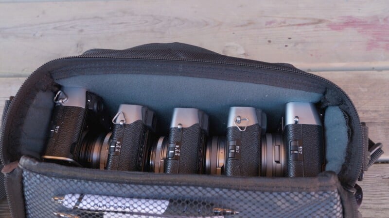 An open black camera bag containing six cameras, all placed side by side. Only the top portions of the cameras are visible, showcasing their lenses and dials. The bag has a mesh pocket on the inner side of the lid with a pen inside. The background is a wooden surface.