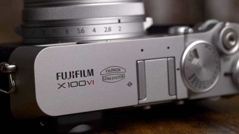 Close-up of a Fujifilm X100V camera, focusing on the branding and dials. The silver body features the "Fujifilm X100V" and "Fujinon Lens System" labels. The image highlights the camera's sleek design and detailed craftsmanship.