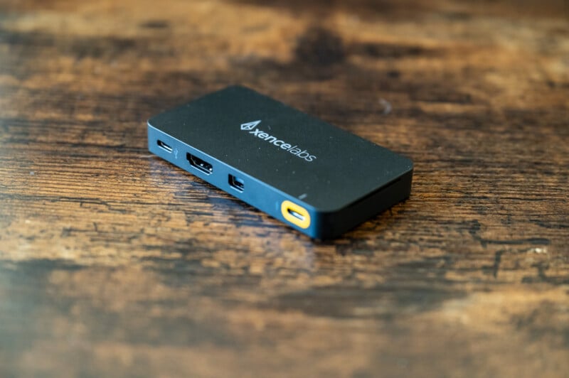 A small, rectangular black device with the "Xencelabs" logo on top is placed on a wooden surface. The device has various ports on its sides, including USB and HDMI connectors. The surface shows a weathered texture with different shades of brown.