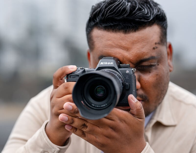 A person with dark, wavy hair and a trimmed beard, wearing a beige shirt, is holding a Nikon camera close to their face, peering through the viewfinder, preparing to take a photograph. The background is blurred, putting the focus on the person and the camera.