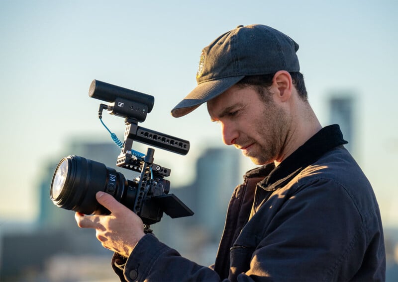 A man wearing a cap and jacket is closely examining a professional camera rig outdoors. He appears focused on the camera’s settings. The background is slightly blurred but shows some tall buildings, suggesting an urban setting. The lighting is natural, from the sun.
