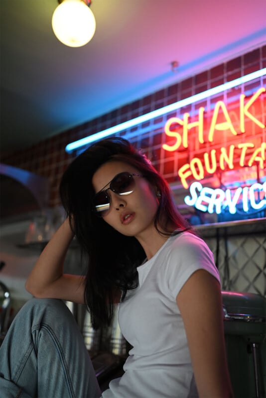 A woman with long black hair is sitting in a retro diner, wearing dark sunglasses, a white t-shirt, and blue jeans. Behind her, a colorful neon sign reads "SHAK FOUNTA SERVICE" against a tiled wall. The scene is dimly lit with a warm, nostalgic ambiance.