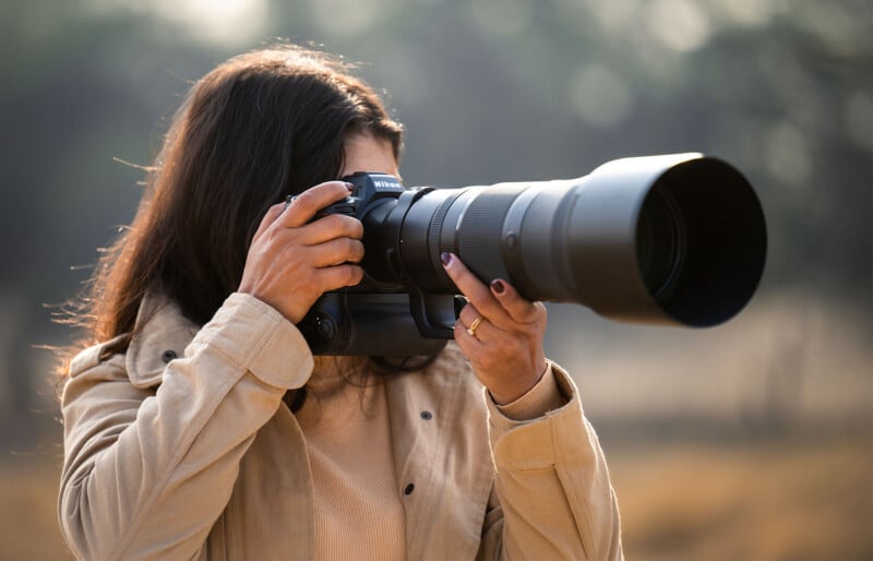 A person with long dark hair, wearing a beige jacket, is holding a large DSLR camera with a long telephoto lens outdoors. They appear to be focusing on capturing a photo, and the background is blurred with natural light filtering through.