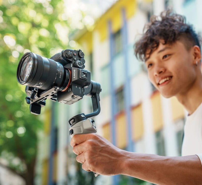 A person with curly hair smiles while holding a professional camera mounted on a handheld stabilizer. They appear to be outdoors, with a colorful building and greenery blurred in the background.